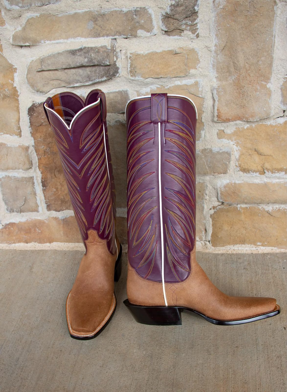 Tall plum-colored boot tops with fanned feather stitching and white side piping over tan roughout vamps, photographed against a stone wall